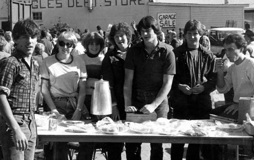 A group of young people at a garage sale table with a crowd behind them and a 'Garage Sale' sign.